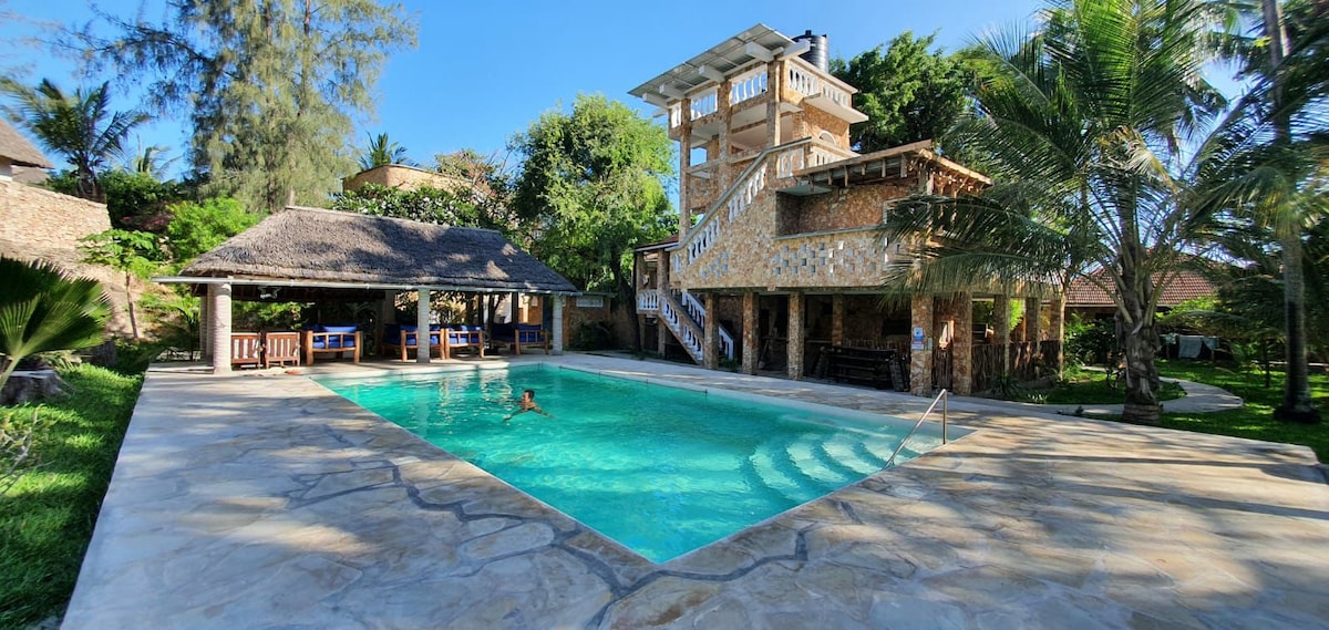 A tranquil pool area is surrounded by lush greenery and tropical plants. Sun loungers are positioned near the water, and a shaded pavilion with seating is visible. A multi-level building stands in the background, enhancing the inviting ambiance of the outdoor space.