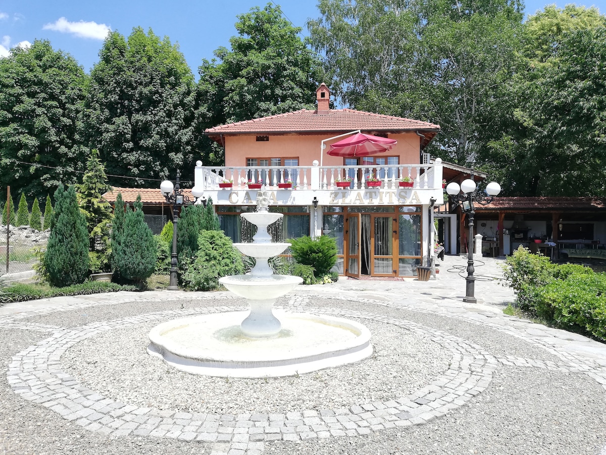 The property features a central fountain surrounded by a landscaped garden. A two-story building with a balcony overlooks the area, partially shaded by a red umbrella. Lush trees line the sides, creating a serene outdoor environment.