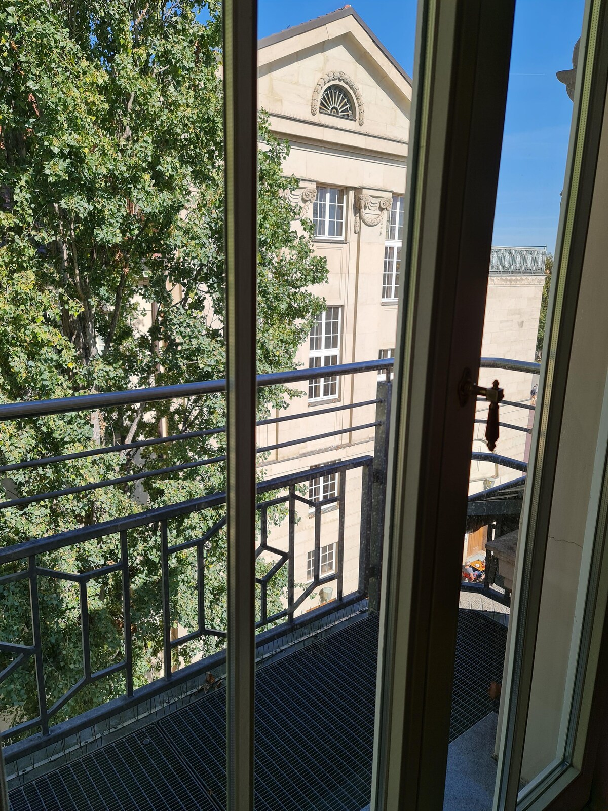 A balcony view is presented, framed by sliding glass doors. Below, a patio is visible, complemented by greenery from a nearby tree. The architectural details of the neighboring building can be seen in the background, showcasing a blend of classic and functional design.