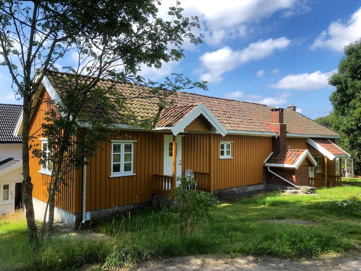 A traditional, renovated wooden house is positioned among greenery, featuring a striking orange facade with white trim. The roof displays a blend of tiles, and windows are adorned with small shutters. A wooden porch is visible at the entrance, providing access to the home.