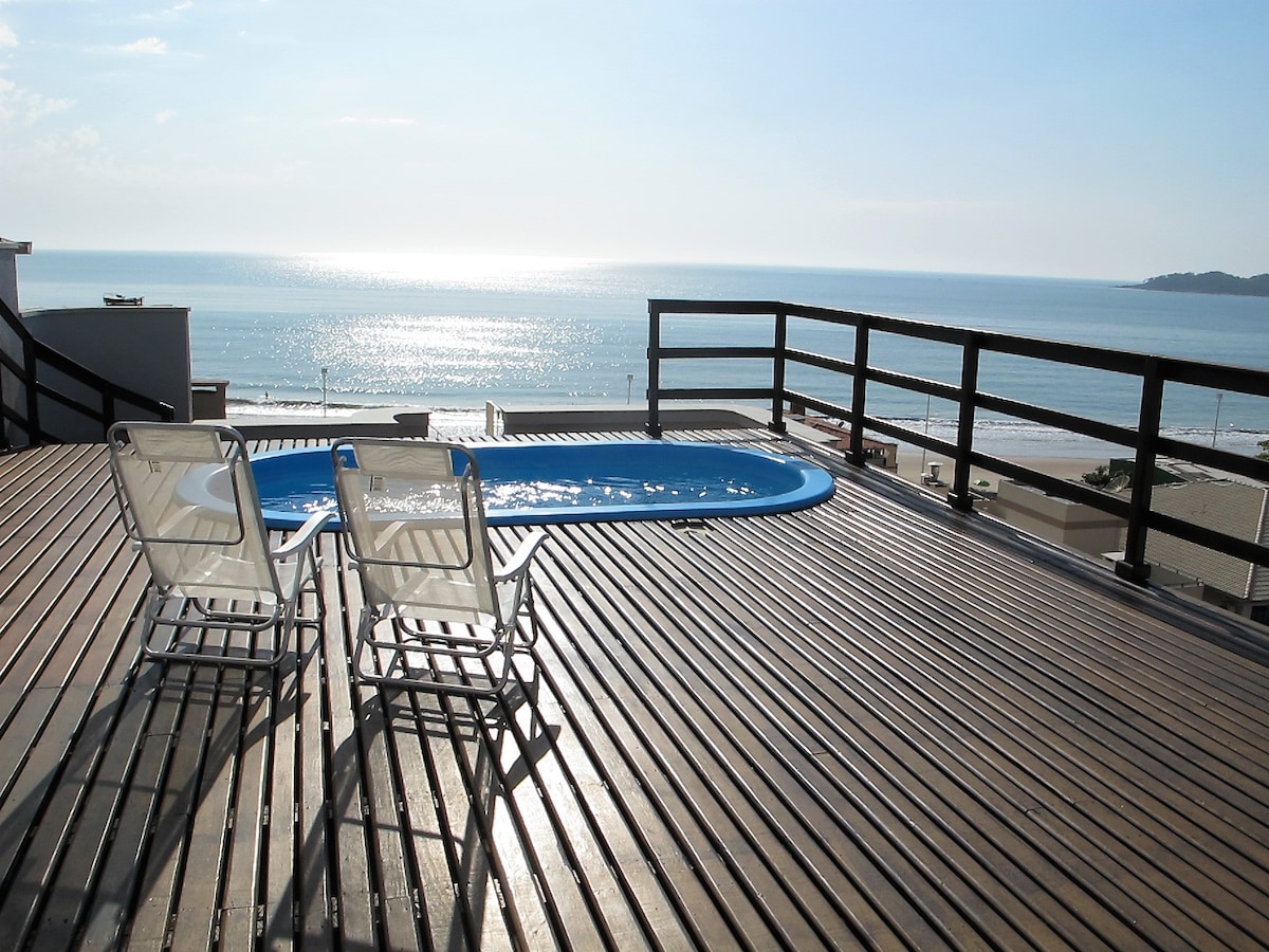 The deck area features a private pool surrounded by wooden flooring, with two white lounge chairs positioned for relaxation. A panoramic view of the ocean sparkles in the sunlight, creating a serene atmosphere. The railings provide safety while framing the scenic landscape.