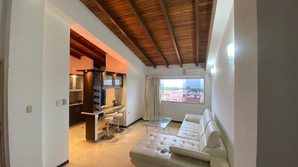A modern living area features a sleek white sofa and a glass coffee table. A work desk with a chair is positioned beside a large window, allowing natural light to fill the space. The high wooden ceiling adds an open, airy feel.