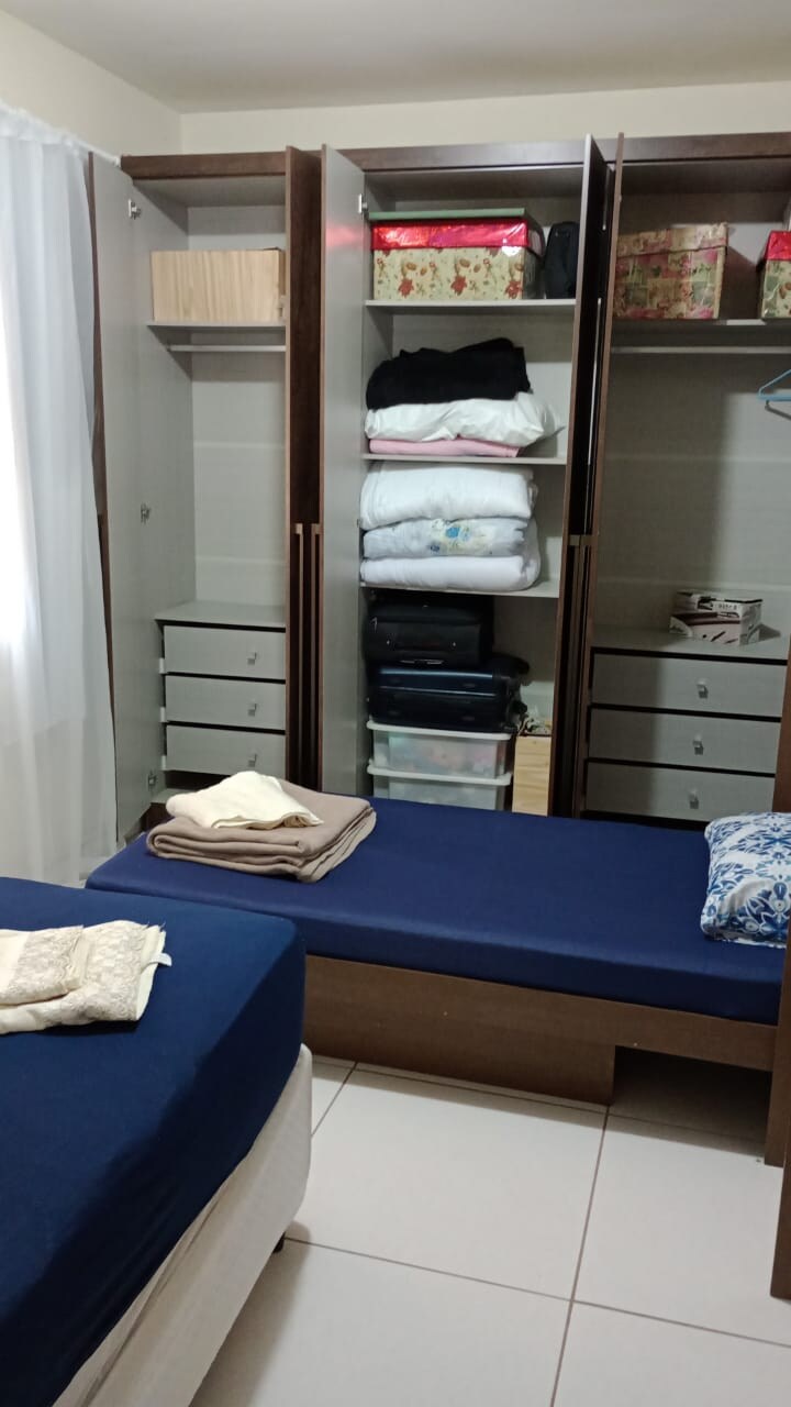A bedroom is featured with two separate beds dressed in dark blue bedding. An organized wardrobe is shown, with shelves displaying neatly stacked blankets and boxes. Drawer units at the bottom offer additional storage, and natural light enters through a nearby window covered by light curtains.