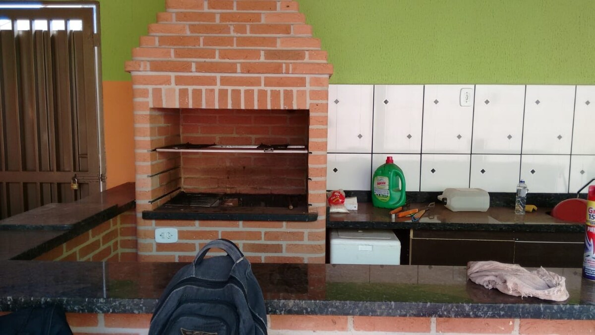 A brick barbecue grill is situated in the kitchen area, accompanied by a stone countertop. The wall features white tiles with black accents. Cleaning supplies and utensils are visible on the countertop, enhancing the functionality of this communal cooking space.