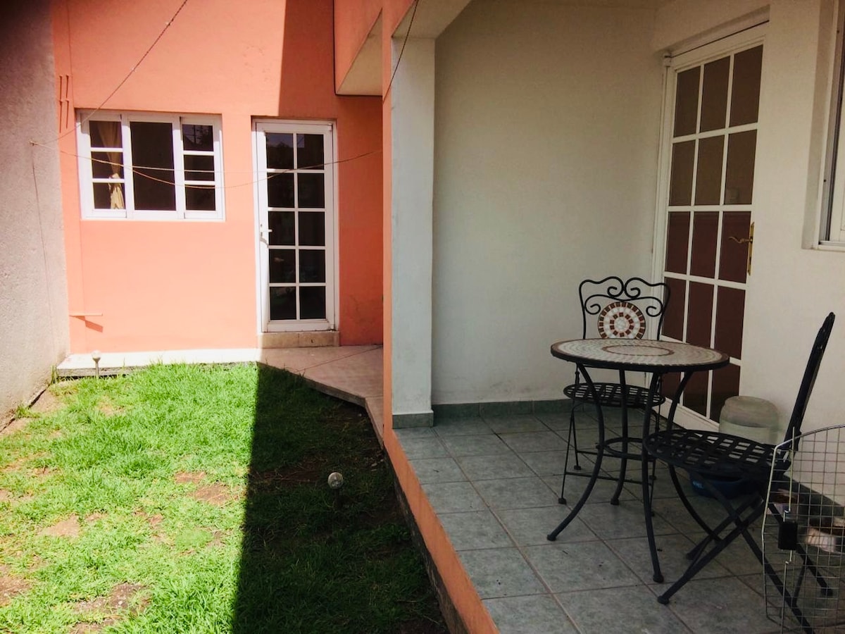 A view of a grassy outdoor area features a small patio with a round table and two chairs. Directly ahead, a door leads into the interior of the house. Light falls across the floor, creating a contrast between shaded and sunny areas.