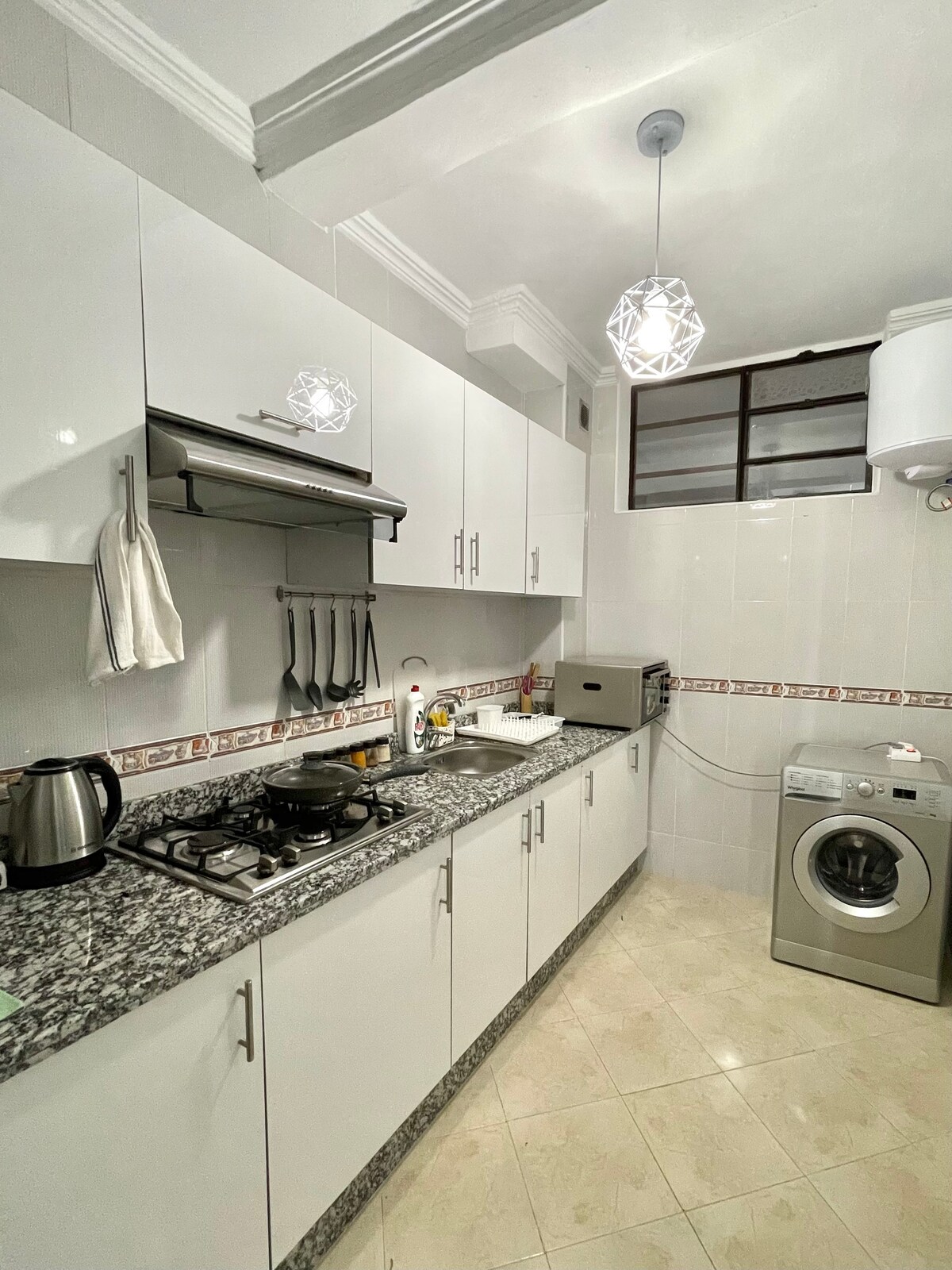 A functional kitchen is presented with sleek white cabinets and a granite countertop. Key appliances include a kettle, a gas stove, and a washing machine. Natural light enters through a window, enhancing the bright and clean environment.
