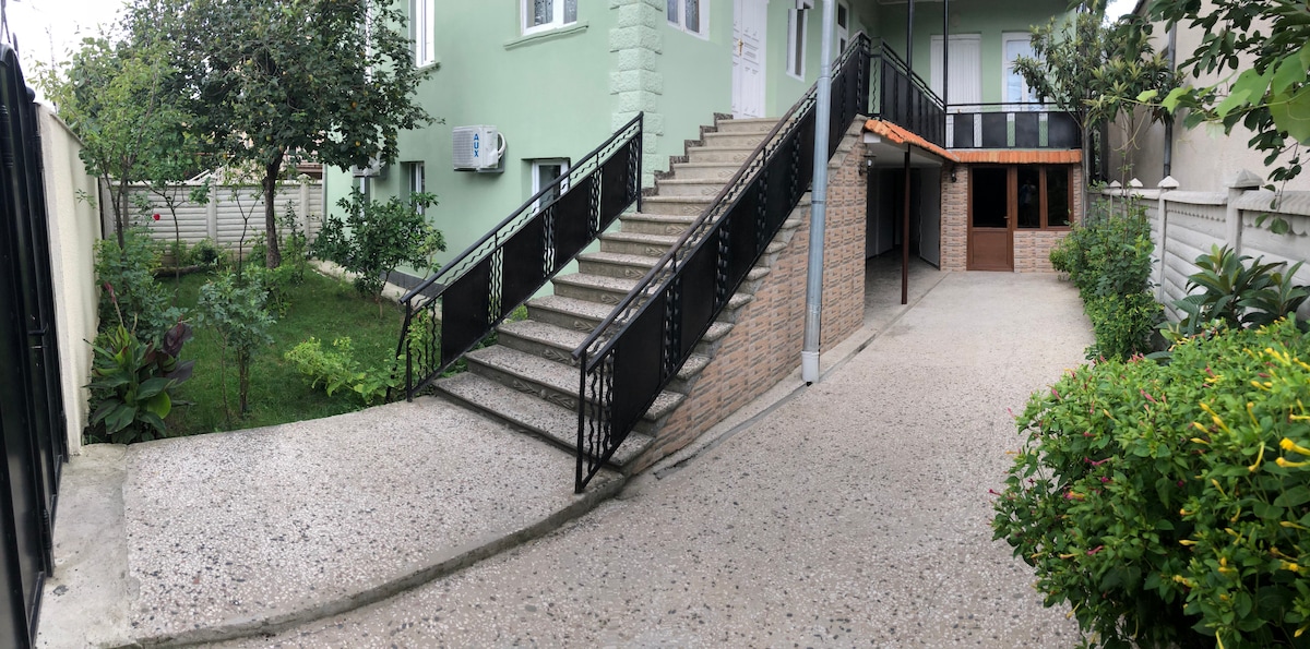 A serene outdoor area is depicted, featuring a winding path leading to a set of stairs. Lush greenery is visible alongside a landscaped garden, while a covered parking space is shown on the right. The stair railing exhibits decorative ironwork, promoting accessibility.