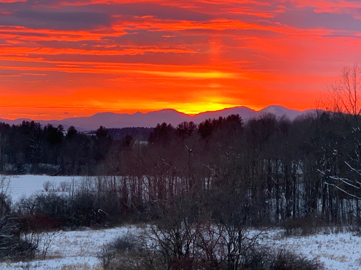 A vibrant sunset casts warm orange and red hues across the sky, with distant mountain silhouettes visible on the horizon. The foreground features a snow-covered landscape dotted with trees, creating a serene winter scene.