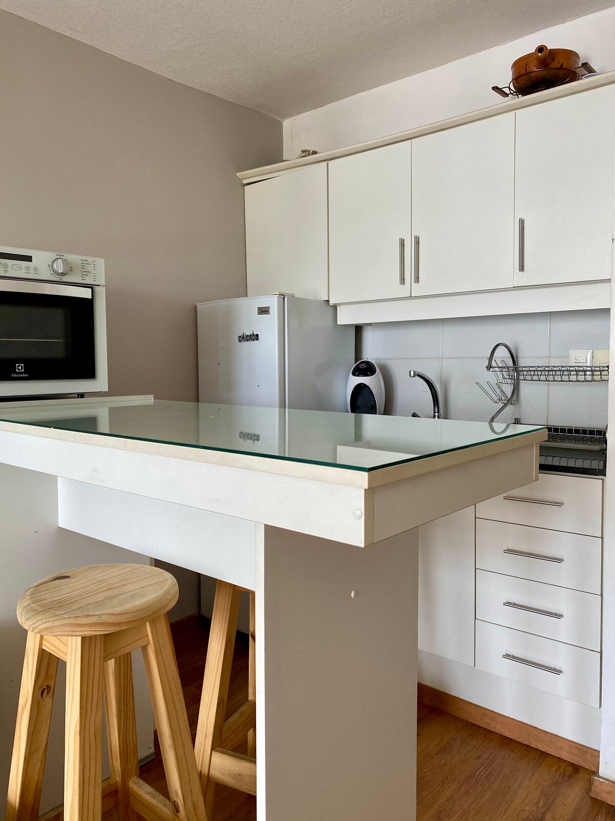 A modern kitchen features sleek white cabinetry and a minimalist glass-top table. A wooden stool provides seating. Stainless steel appliances, including a microwave and refrigerator, are visible, alongside a small sink and neatly arranged kitchenware.