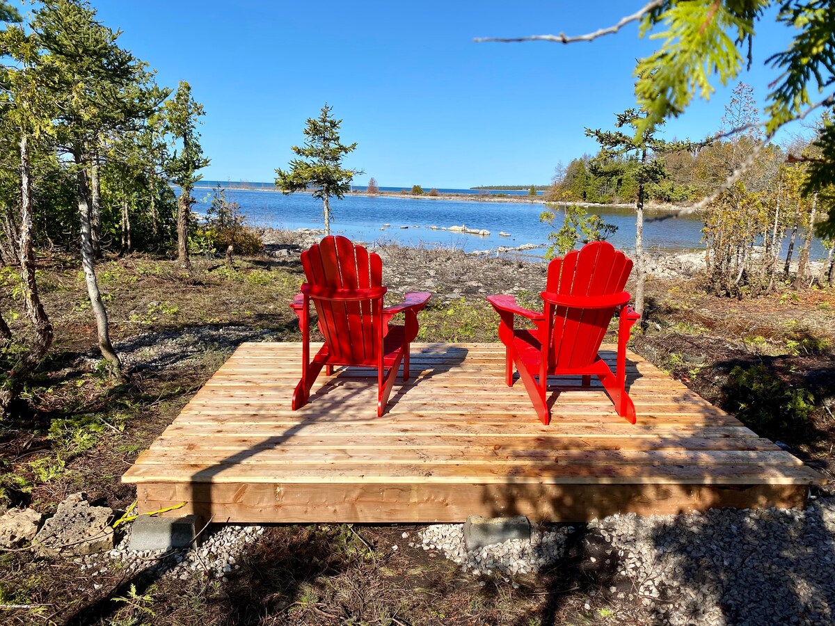 A wooden deck is positioned by the water's edge, featuring two red adirondack chairs facing the serene lake. The surrounding area is filled with small trees and natural landscaping, providing an unobstructed view of the water and sky.