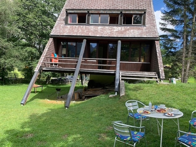 A charming A-frame house is set on a grassy area, featuring large front windows and an elevated deck. Outdoor seating is arranged at a table with colorful chairs, while a grill is positioned nearby, surrounded by trees that provide a natural backdrop.
