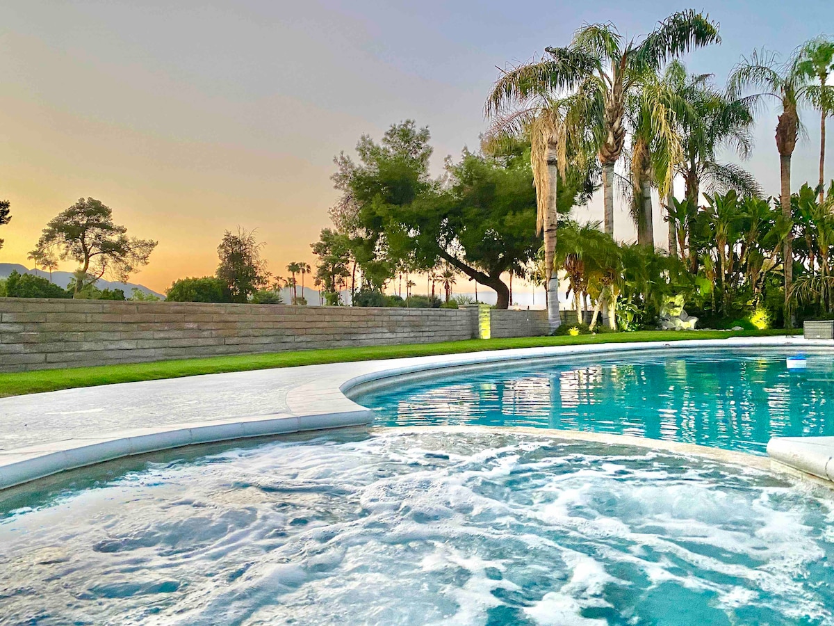 A tranquil view of the pool area showcases clear water with gentle ripples. Lush palm trees and greenery surround the space, while soft sunset hues illuminate the sky in the background, enhancing the serene atmosphere.