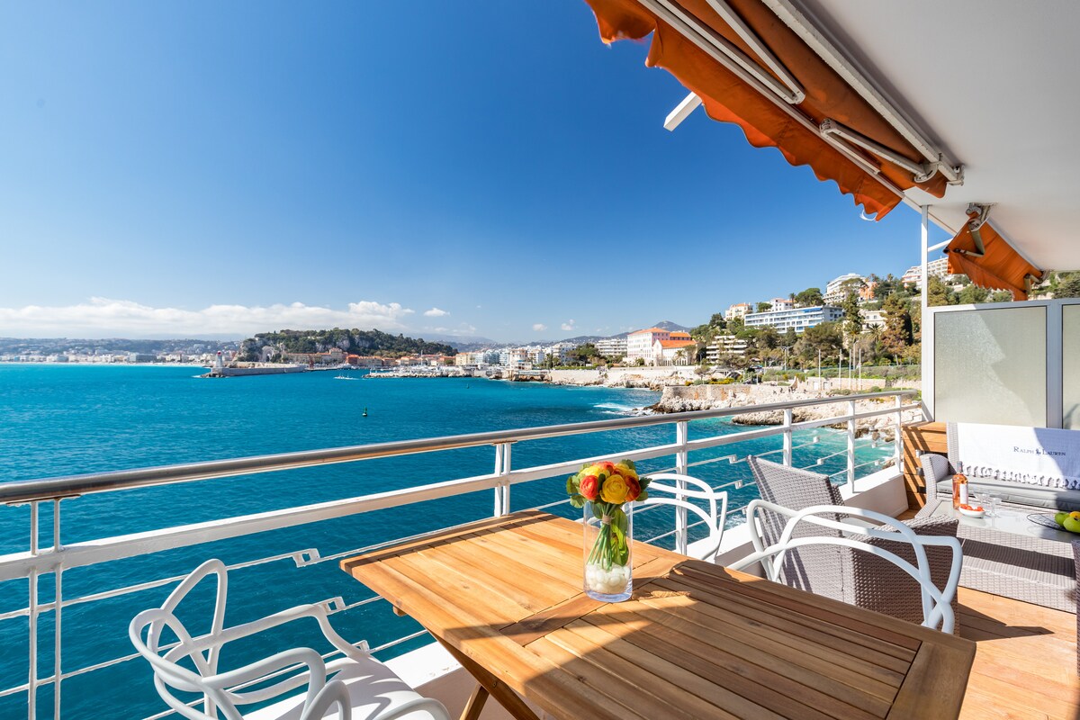 A deep terrace features a wooden table surrounded by four chairs, with a view of the tranquil blue sea and the coastline. Bright sunlight illuminates the space, while a vase of colorful flowers enhances the setting. Awnings provide shade, creating a comfortable outdoor area.