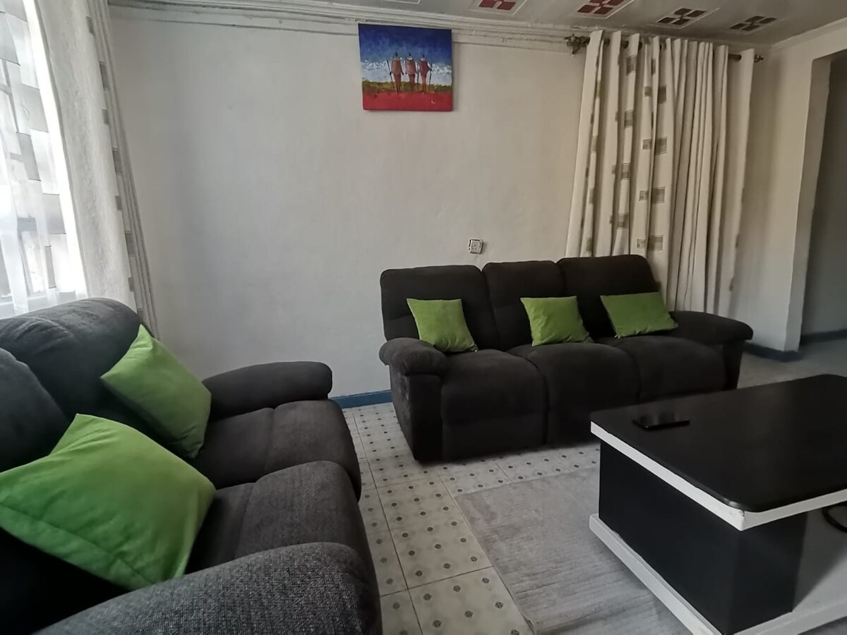 A cozy living area features two dark gray sofas adorned with green cushions, inviting relaxation. Natural light enters through sheer curtains, illuminating the space. A simple coffee table rests in the center, complemented by a light-colored rug. Colorful artwork adds a touch of charm to the wall.