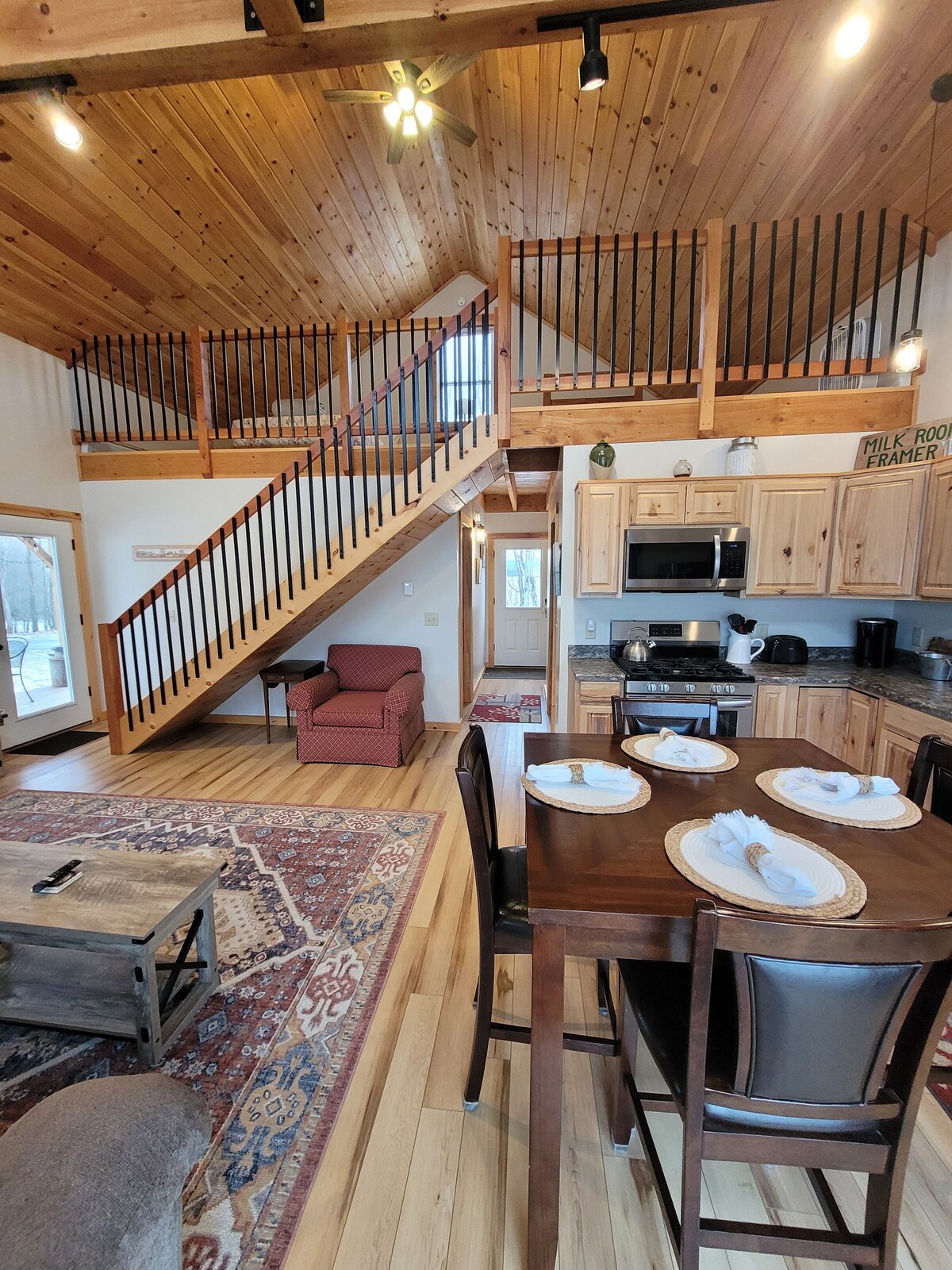 The open kitchen and dining area feature a wooden table with place settings, complemented by a cozy seating nook. Natural light enters through large windows, illuminating the wood-paneled walls and ceiling. A staircase leads to a lofted area, enhancing the spaciousness of the room.