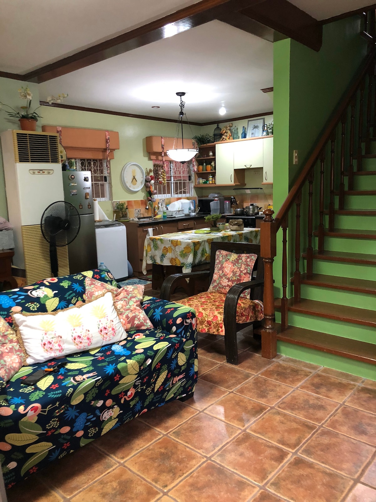 A living area is shown, featuring a colorful patterned couch and a wooden armchair situated on tiled flooring. A staircase with green steps leads to the upper level. The open kitchen is visible in the background, equipped with appliances and kitchenware.