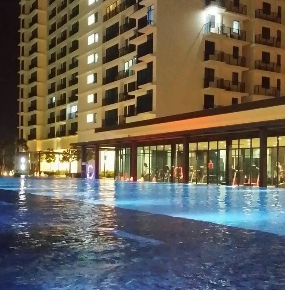 The swimming pool is illuminated at night, showcasing a tranquil blue hue. Surrounding the pool are modern buildings with balconies, and large windows highlight the gym area inside. Soft lighting enhances the peaceful atmosphere, providing a calming retreat.