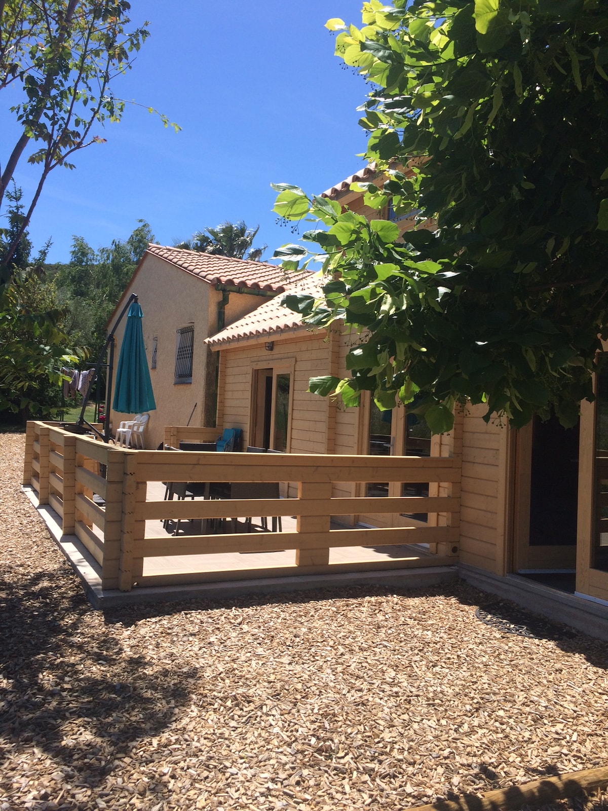 The exterior of the chalet is showcased, featuring a wooden deck surrounded by greenery. A sun umbrella stands above outdoor seating, providing a shaded area for relaxation. The space is framed by sunny blue skies and a neatly maintained garden path.