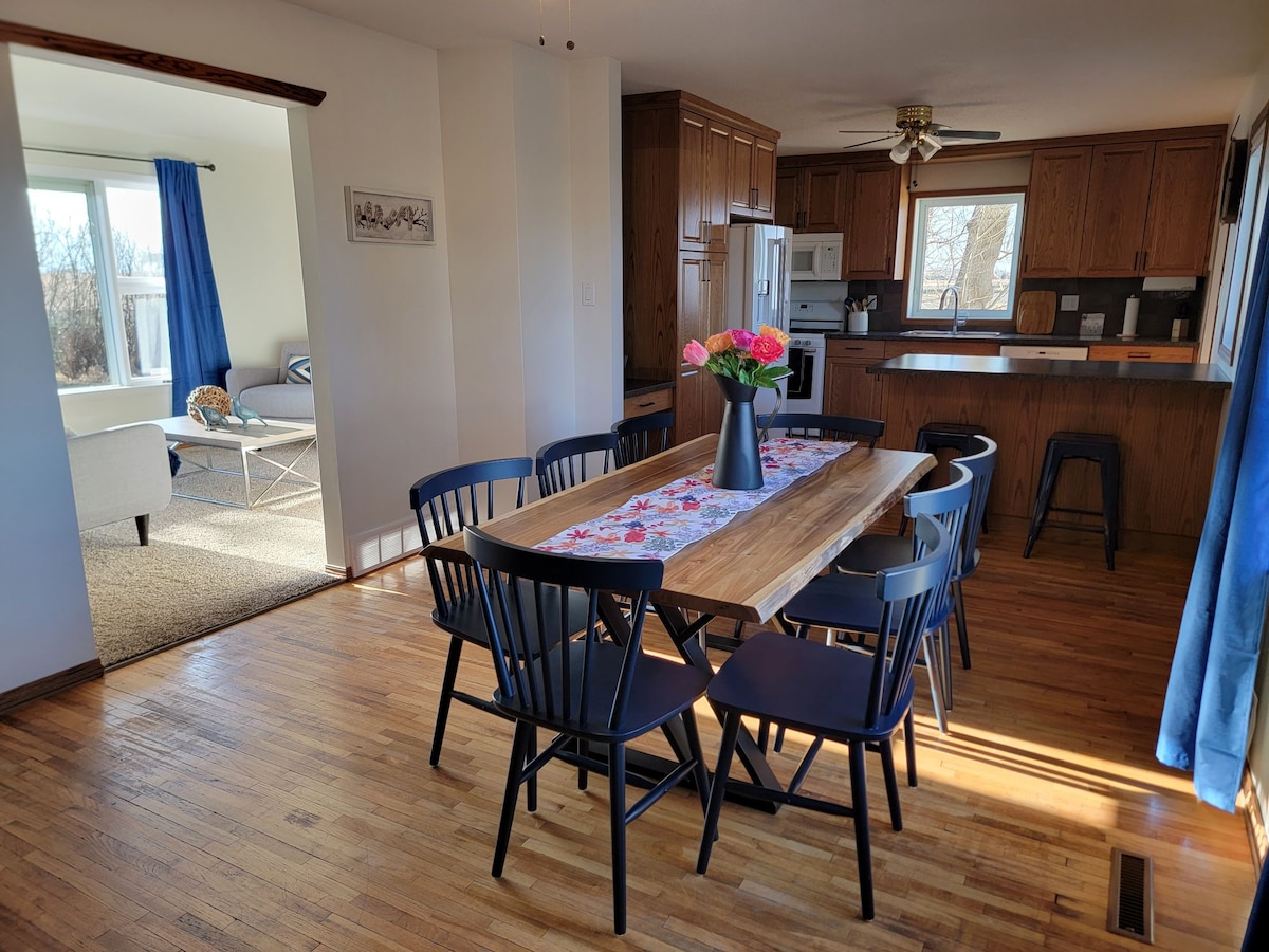 A spacious dining area is highlighted by a long wooden table surrounded by black chairs. A floral centerpiece sits atop the table, while the kitchen features wooden cabinets and modern appliances. Natural light filters in through large windows with blue curtains.