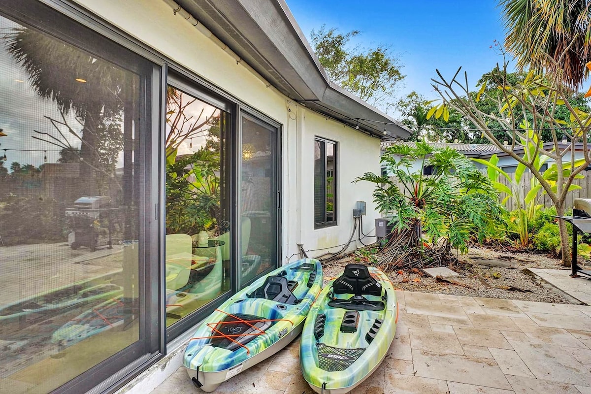 Two kayaks are positioned outside a modern home, resting on a tiled patio. The vibrant kayaks are surrounded by lush greenery, including palm trees and various plants, contributing to a natural and inviting outdoor space.