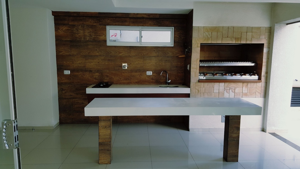 An open kitchen area features a modern white countertop with a wooden base. A wall of natural wood adds warmth, while a built-in grill is visible to the side. Natural light streams through a window, illuminating the sleek space designed for functionality.