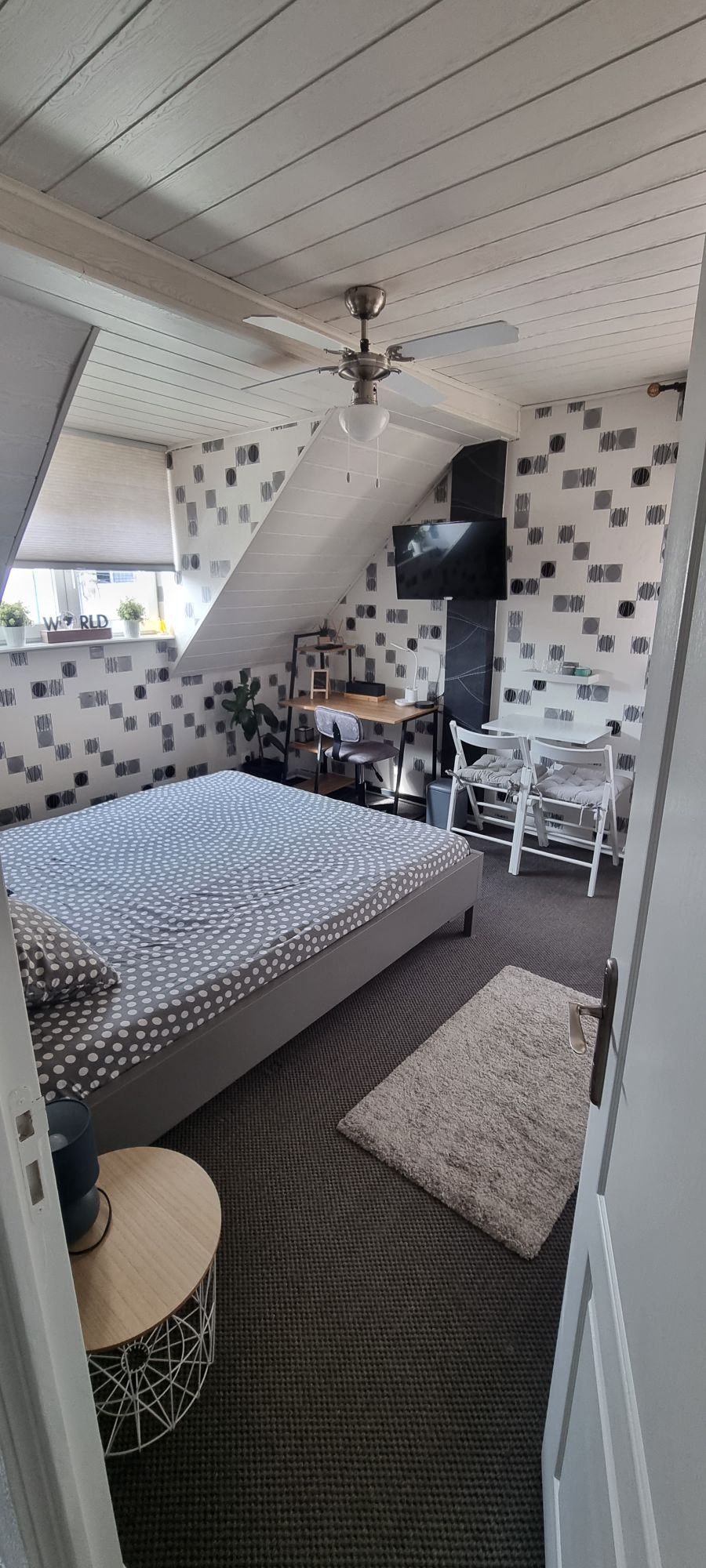 A spacious bedroom is visible, featuring a double bed with a patterned bedspread. A small table and two chairs are arranged near a window, which is adorned with plants. A light-colored rug is placed on the floor, and a ceiling fan is mounted above.