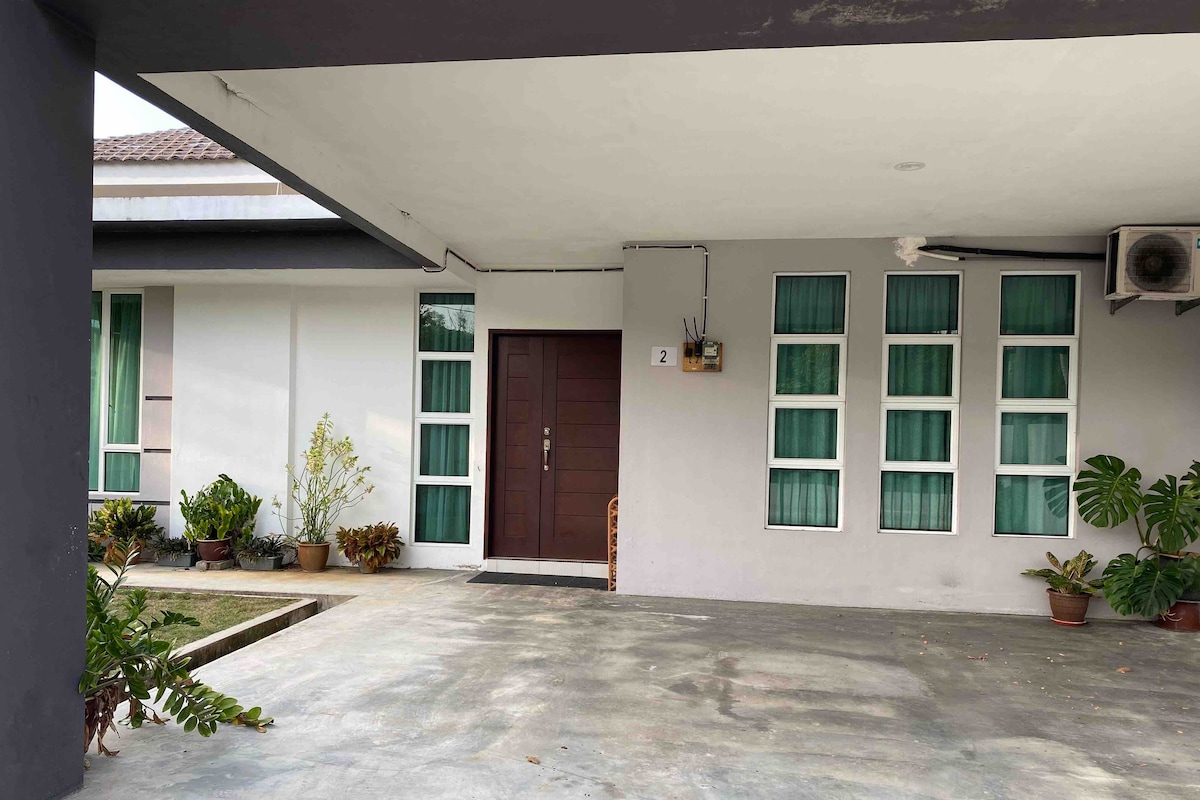 A front view of the homestay features a spacious porch with a concrete floor, bordered by potted plants. The entrance displays a solid brown door, while a series of six rectangular green window frames adorn the right side, allowing natural light to fill the interior.