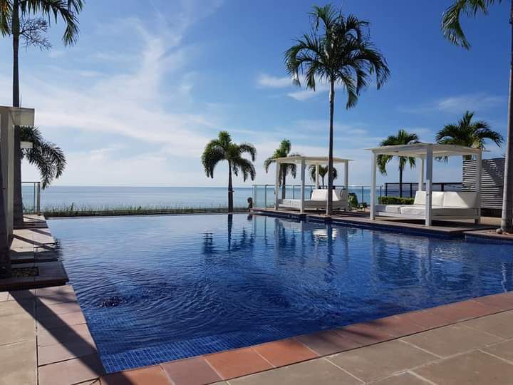 An expansive pool area overlooks a serene body of water. Palm trees gently sway in the breeze, and two shaded cabanas provide comfortable lounging spaces. The clear blue water of the pool reflects the bright sky, creating a calming atmosphere.