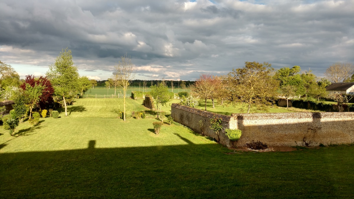 An expansive garden is depicted, featuring manicured grass and a variety of trees and shrubs. Stone walls enclose the garden, providing privacy. In the distance, fields stretch under a cloudy sky, adding depth to the serene outdoor setting.