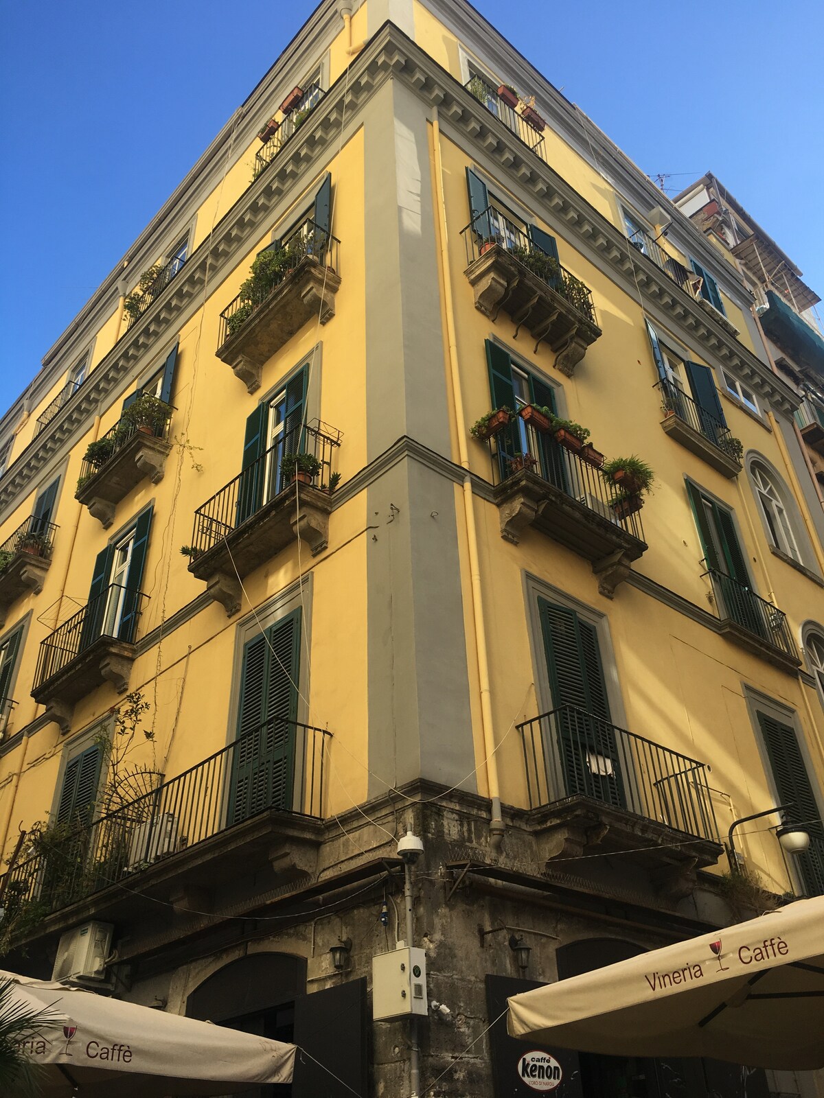 The exterior of a historic building is showcased, featuring a yellow façade adorned with green shutters. Balconies with potted plants extend outward, creating a charming urban profile. The image captures the architectural details and vibrant colori that characterize the lively atmosphere of the neighborhood.