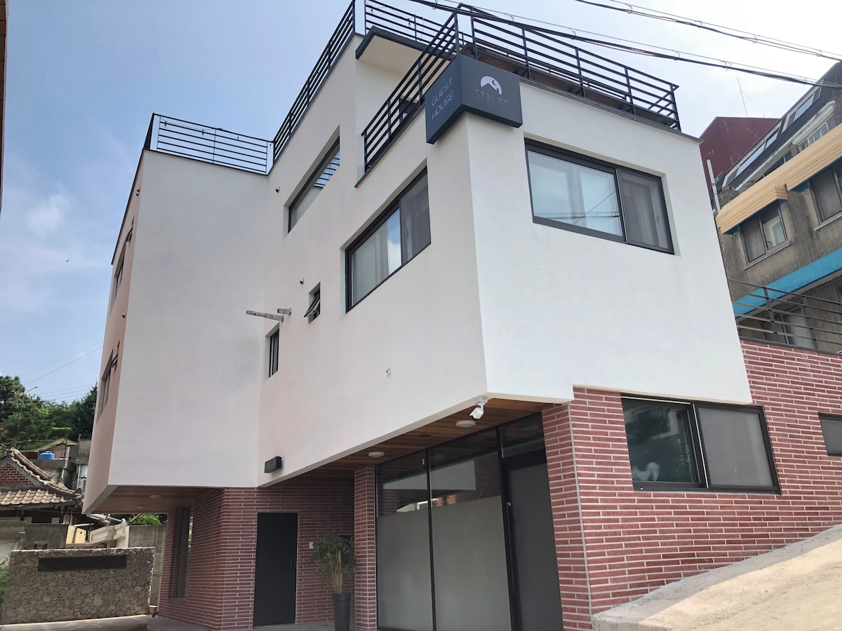 A modern three-story guesthouse is displayed, featuring a combination of white and brick exterior walls. Large windows allow natural light into the space, while a rooftop terrace is accessible, providing additional outdoor space.