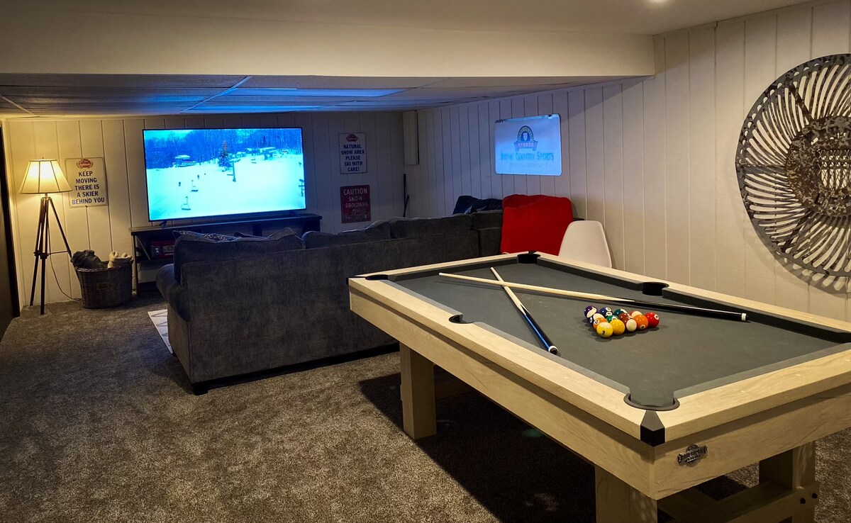 A spacious finished basement features a billiards table in the foreground, with a comfortable sectional couch nearby. A large television displays a winter scene, providing entertainment options. Soft lighting creates a relaxed environment, while carpeted flooring adds warmth.