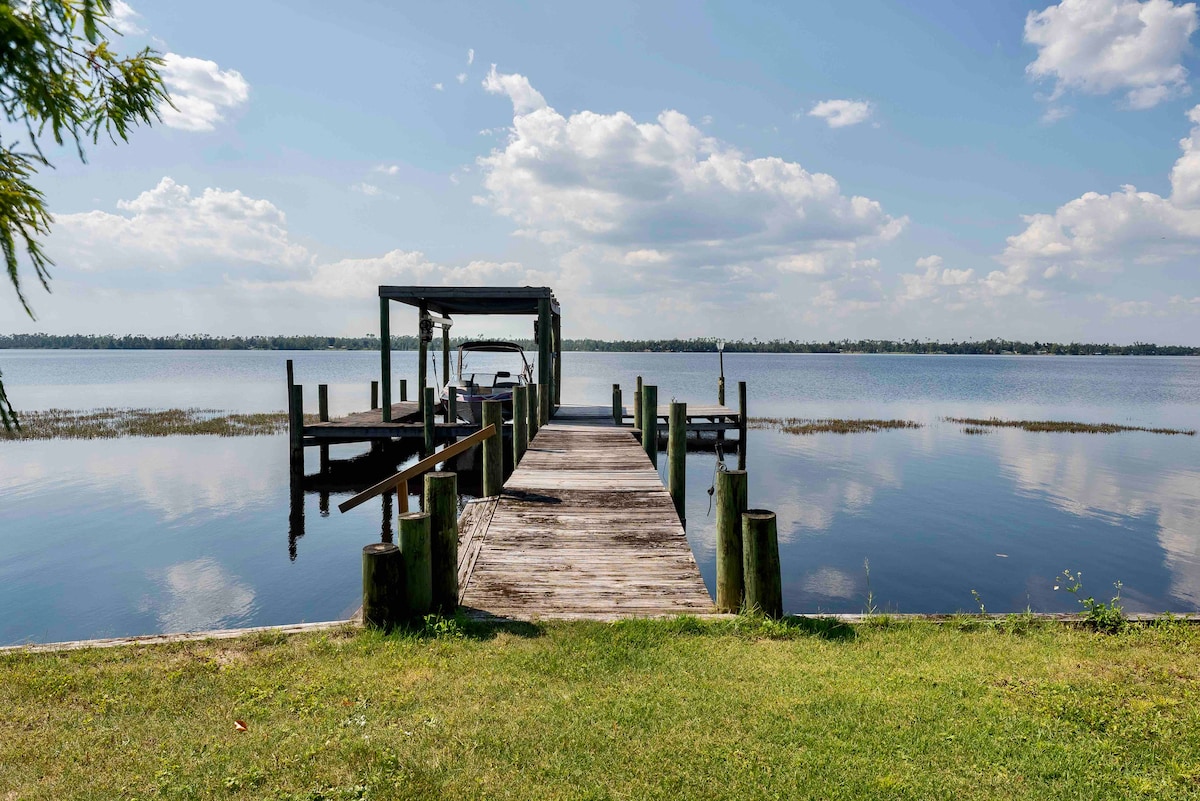 A wooden dock extends over calm waters, leading to a covered area for boats. Lush green grass frames the dock, while puffy clouds drift above a serene lake, creating a peaceful connection with nature.