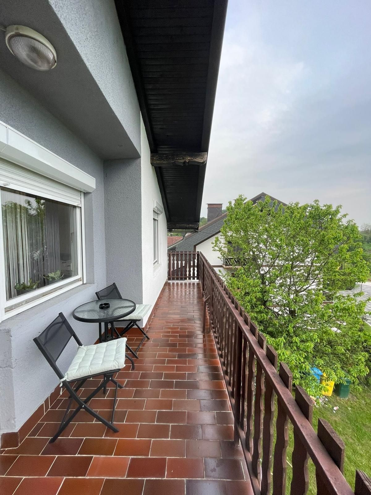 A balcony is shown with tiled flooring and a wooden railing. Two metal chairs with cushions and a small round table occupy the space, providing a view of surrounding greenery and nearby structures.