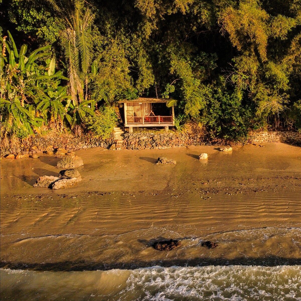 A secluded beach area features a wooden structure set against lush greenery. The structure is positioned above the sandy shore, where water gently laps at the rocks. Vibrant plants surround the area, adding to the natural feel of this tranquil setting.