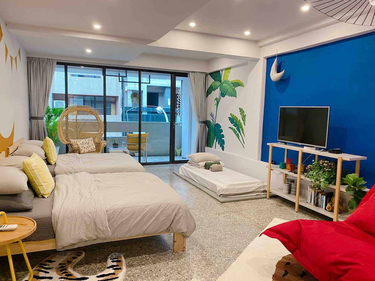 A spacious room features two beds with light linens, complemented by a colorful accent wall. A large window allows natural light, providing views of an outdoor area. A seating space includes a cozy chair, and a television is mounted on a wooden stand with plants arranged nearby.