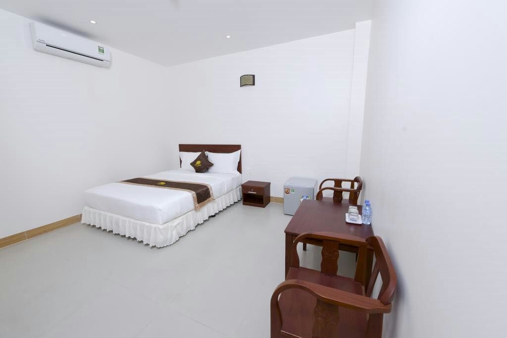 A bright room features a spacious bed with a white bedspread and decorative pillow. A small desk and two chairs are positioned near a mini fridge, while an air conditioning unit is mounted on the wall for comfort.