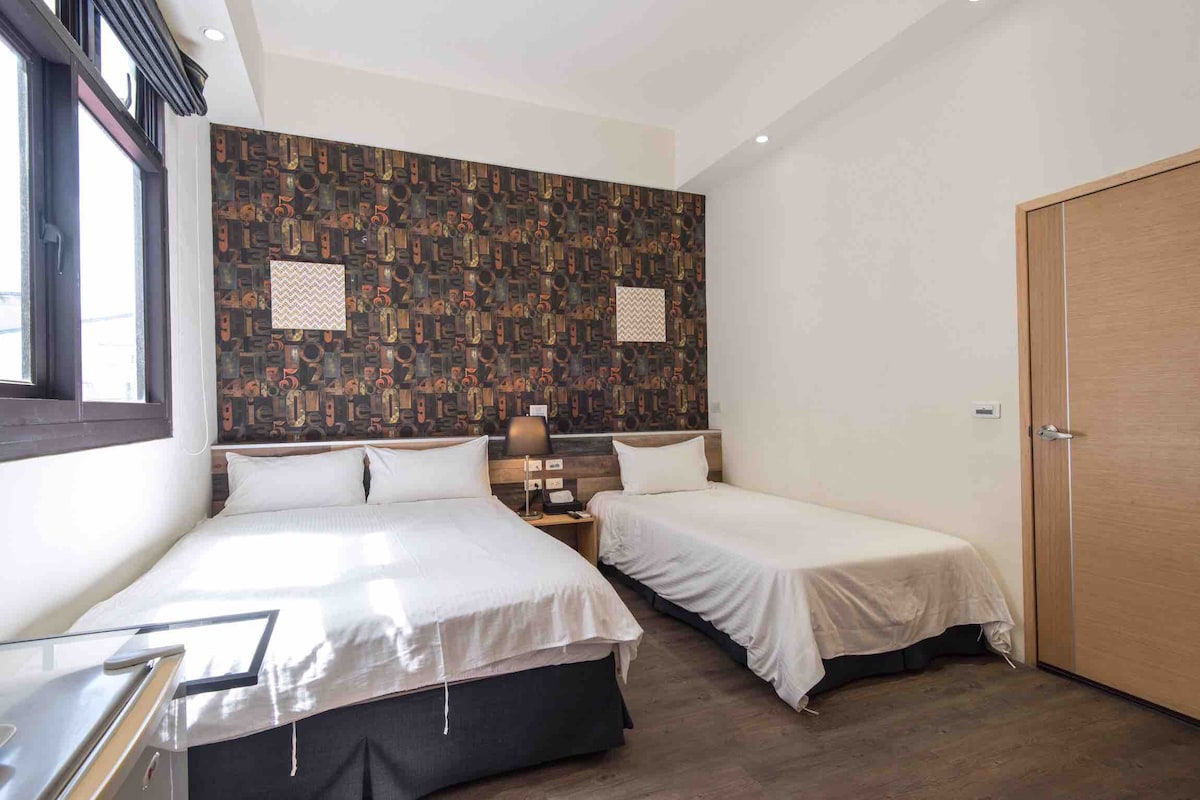 A spacious room features two beds, one with a white bedspread and the other with a light grey covering. A decorative wallpaper adds texture to the wall behind the beds. Natural light enters through a large window, and two bedside tables are positioned beside the beds.