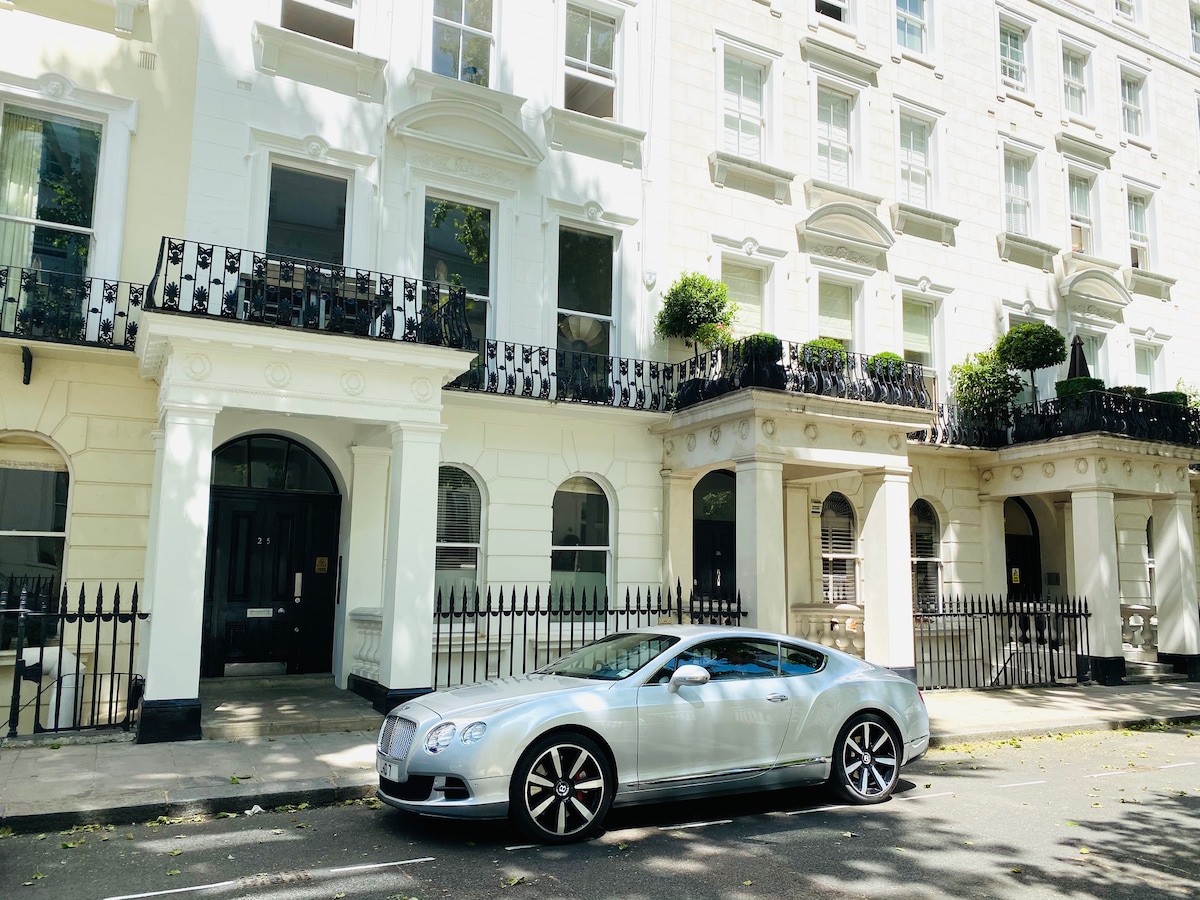A classic white building is depicted, showcasing elegant architecture and prominent arched windows. A silver car is parked in front, complementing the facade. The entrance features decorative iron railings and potted greenery, contributing to the refined atmosphere of the property.