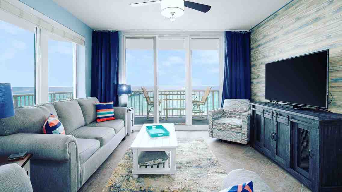 A bright living area features large sliding glass doors leading to a balcony, allowing for panoramic views of the coast. Comfortable seating arrangements include a gray sofa and patterned armchair. A flat-screen TV is mounted on a dark wooden cabinet, with light-colored decor enhancing the ambiance.
