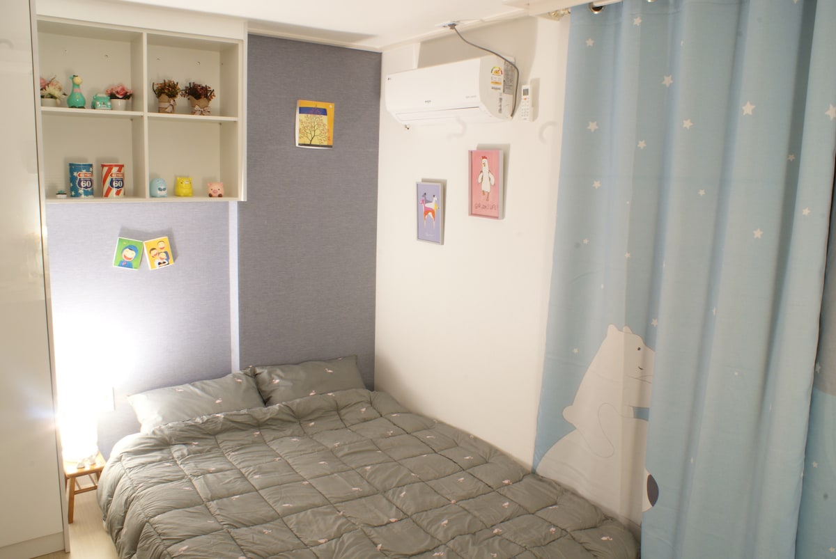 The sleeping area includes a spacious bed dressed in a gray quilt. Soft lighting is provided by a lamp placed on a small bedside table. The wall features cheerful artwork and decorative shelves filled with small items, alongside a light blue curtain adorned with playful motifs.
