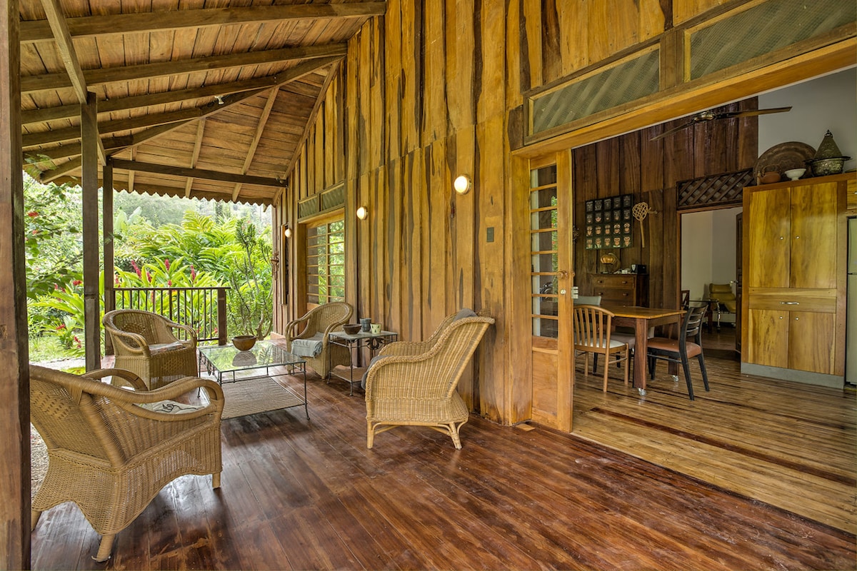 A covered veranda features several rattan chairs and a small coffee table, set against wooden walls and flooring. Large windows open to a lush tropical garden, enhancing the natural ambiance. The inviting space is illuminated by wall-mounted lights, creating a relaxed atmosphere.