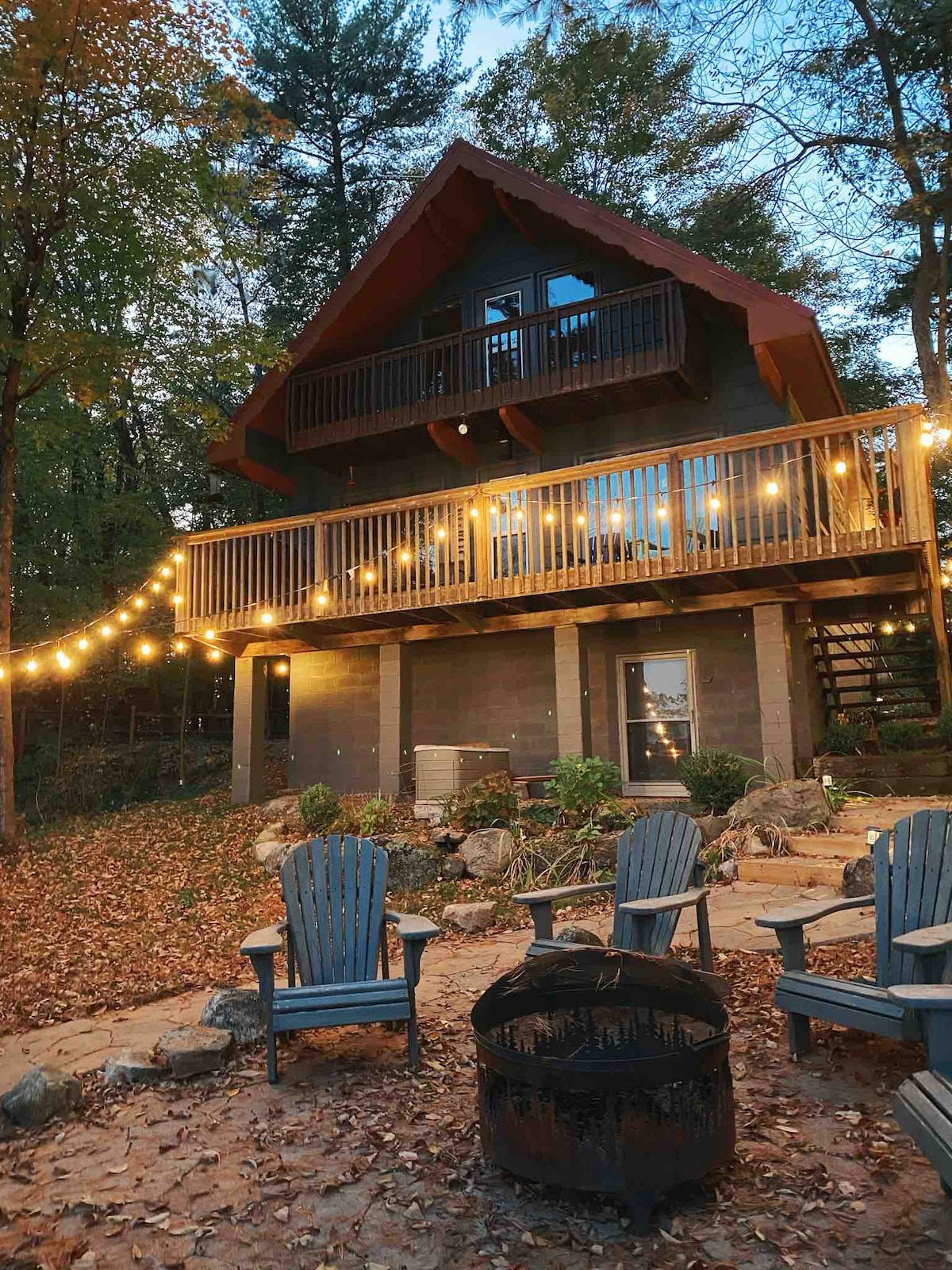 A charming cottage is showcased amid a serene outdoor setting, surrounded by trees and scattered autumn leaves. The balcony, adorned with string lights, overlooks a firepit area with wooden chairs. The inviting facade highlights a unique architectural design, creating a cozy atmosphere.