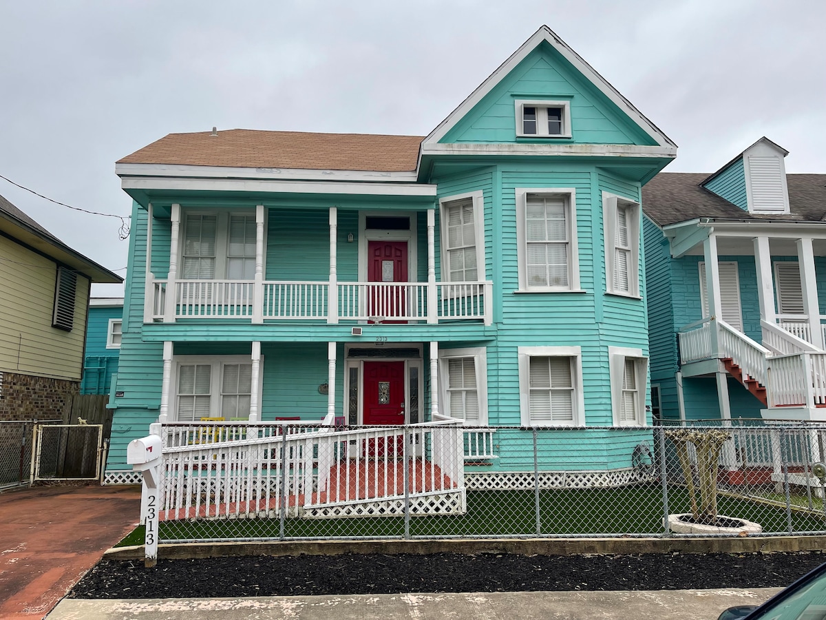 This charming two-story home features a teal exterior with white trim and spacious front porches. The property is bordered by a low fence, and a concrete driveway leads up to the front entrance adorned with a red door.