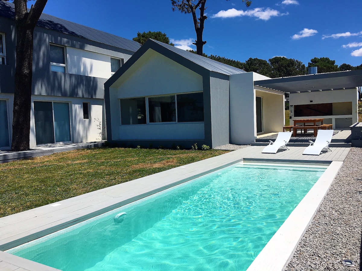 A modern house features a minimalistic design with large windows and a sloped roof. A private swimming pool, surrounded by a wooden deck, glistens under the sunlight. Two lounge chairs are positioned poolside, offering a relaxing area for guests. Lush green grass adds to the tranquil setting.