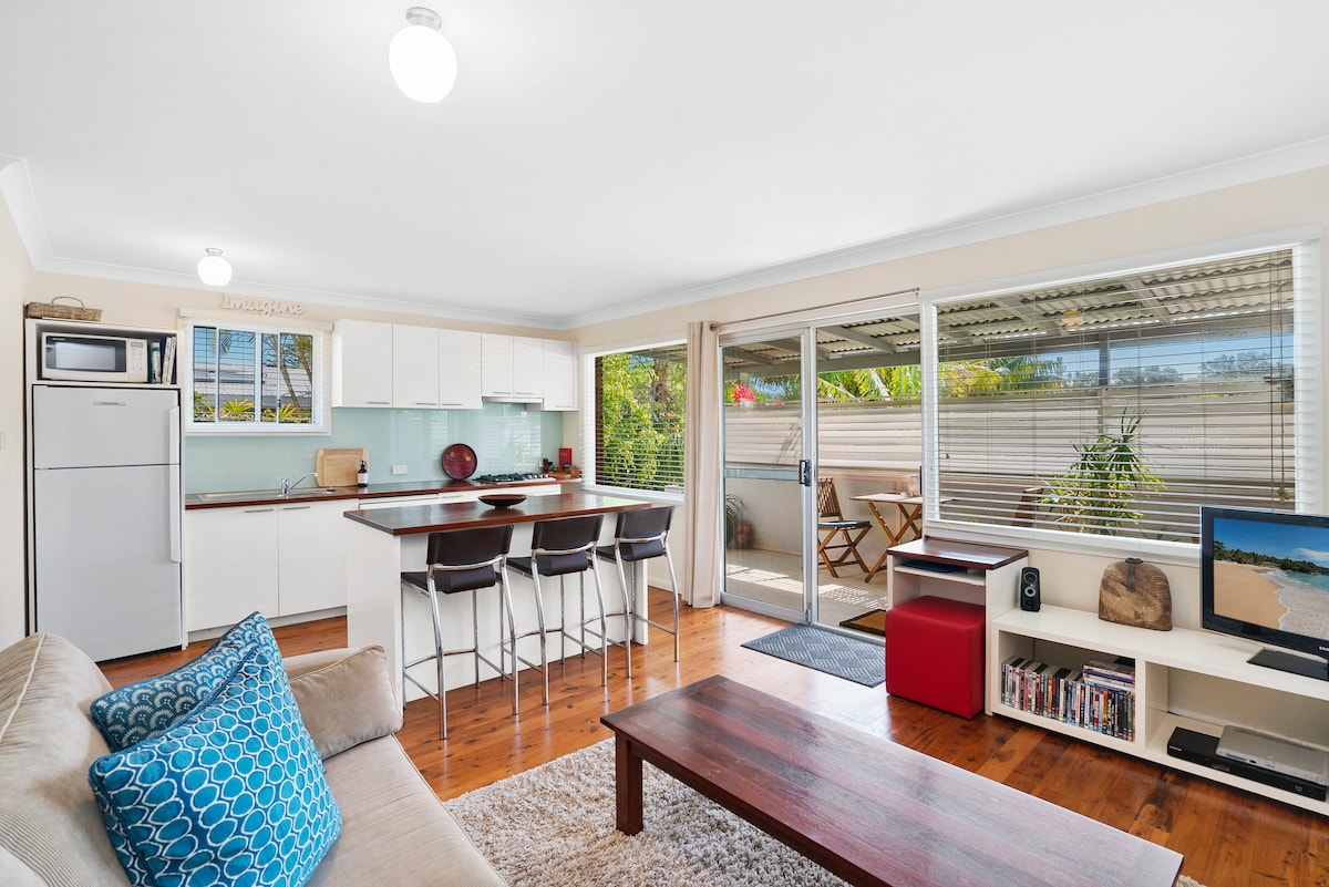 A modern kitchen area is featured with white cabinetry and a counter accommodating four stools. A cozy living space includes a sofa adorned with a blue decorative pillow and a wooden coffee table. Large sliding doors provide access to a small outdoor area with greenery.