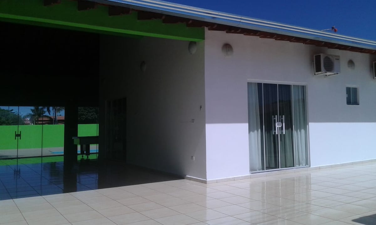 The image reveals a modern home exterior with a bright green accent wall. Large glass doors provide views into a spacious living area, while shiny tile floors reflect natural light. The sky above is clear, complementing the inviting atmosphere of the surrounding outdoor space.