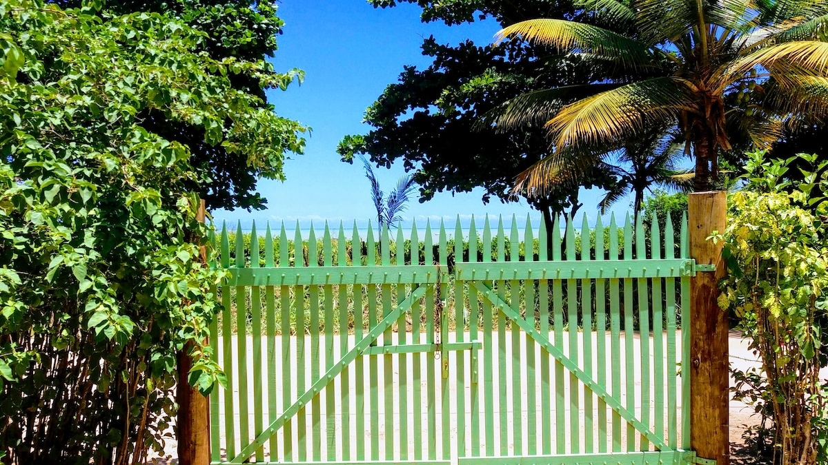 A green, wooden gate stands open, framed by lush greenery and palm trees. In the background, the tranquil blue sea is visible, highlighting the welcoming access to the beach. The clear sky enhances the serene atmosphere of the location.