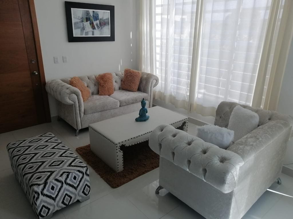 A bright living space features a plush white sofa and armchair, accented with orange cushions. A patterned ottoman adds visual interest, while a decorative table with a blue sculpture serves as a focal point. Natural light filters through sheer curtains.