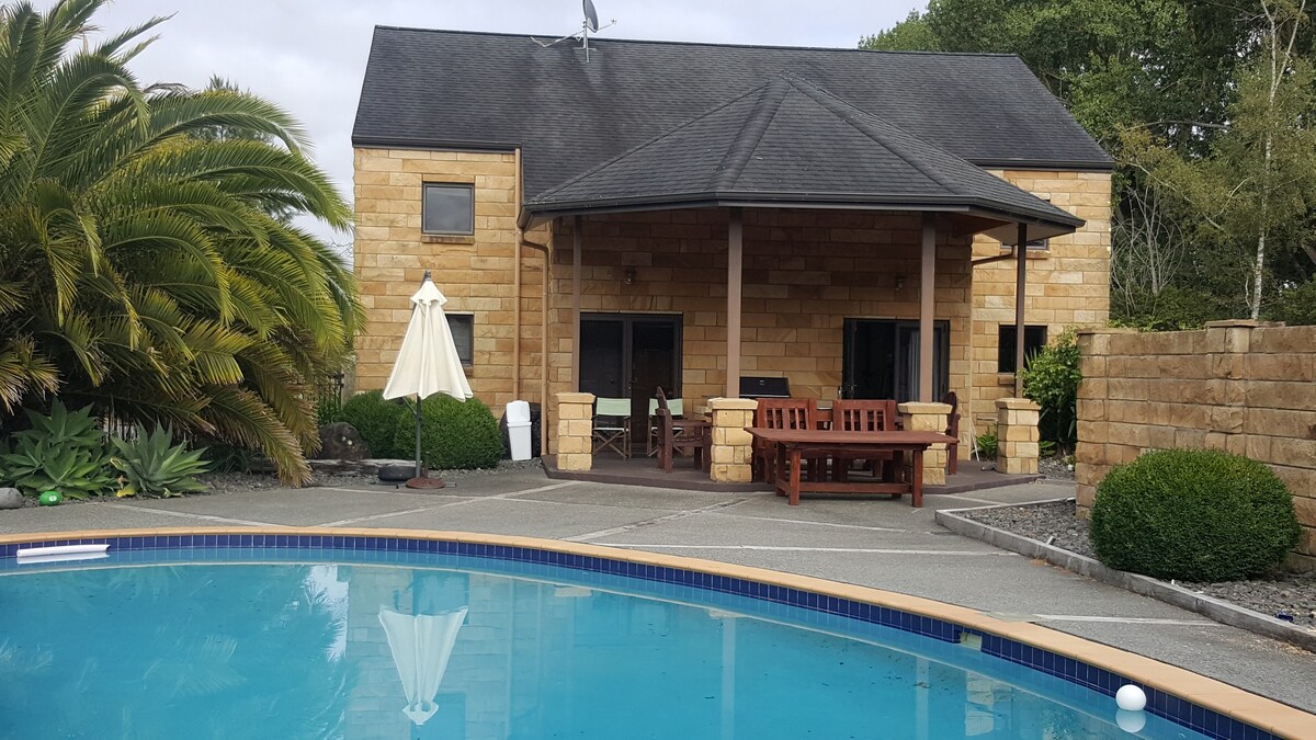 A spacious studio unit is shown, featuring a stone exterior with large glass doors facing a resort-style pool. Surrounding greenery includes tall palm trees and well-maintained shrubs. A wooden dining table and umbrella are situated on the patio, providing an area for relaxation.